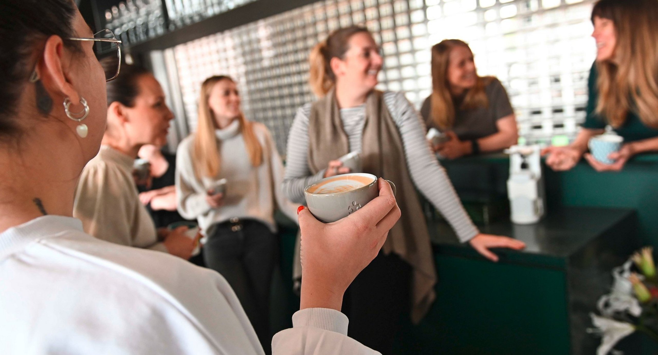 Upart Mitarbeiterinnen stehen mit Kaffee zusammen an der Bar, im Vordergrund Hand mit Kaffeetasse, Hintergrund verschwommen