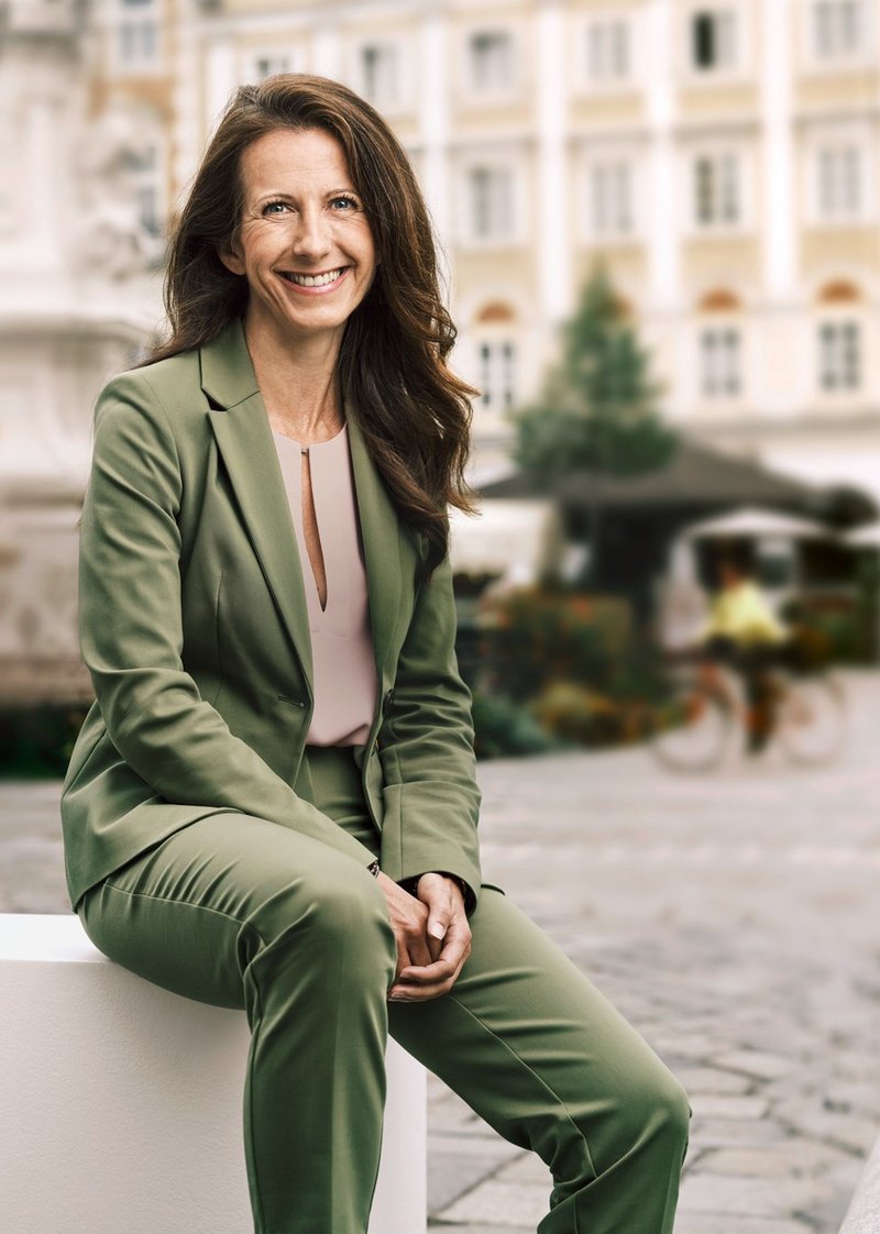 Frau mit braunen Haaren im Olive-Grünem Anzug sitzt am Linzer Hauptplatz
