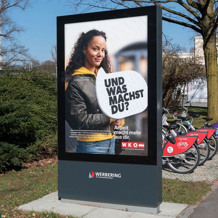 Citylight Plakat, Kampagne der WKO Ö, junges Mädchen steht bei Straßenbahnhaltestelle, mit Sprechblase, "... und was machst du?"