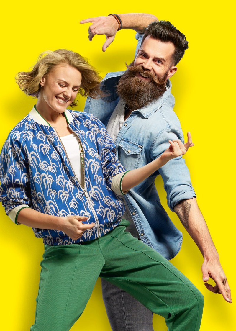 gelber Hintergrund, Frau im Vordergrund, spielt Luftgitarre und Mann mit Bart steht tanzend hinter der Frau